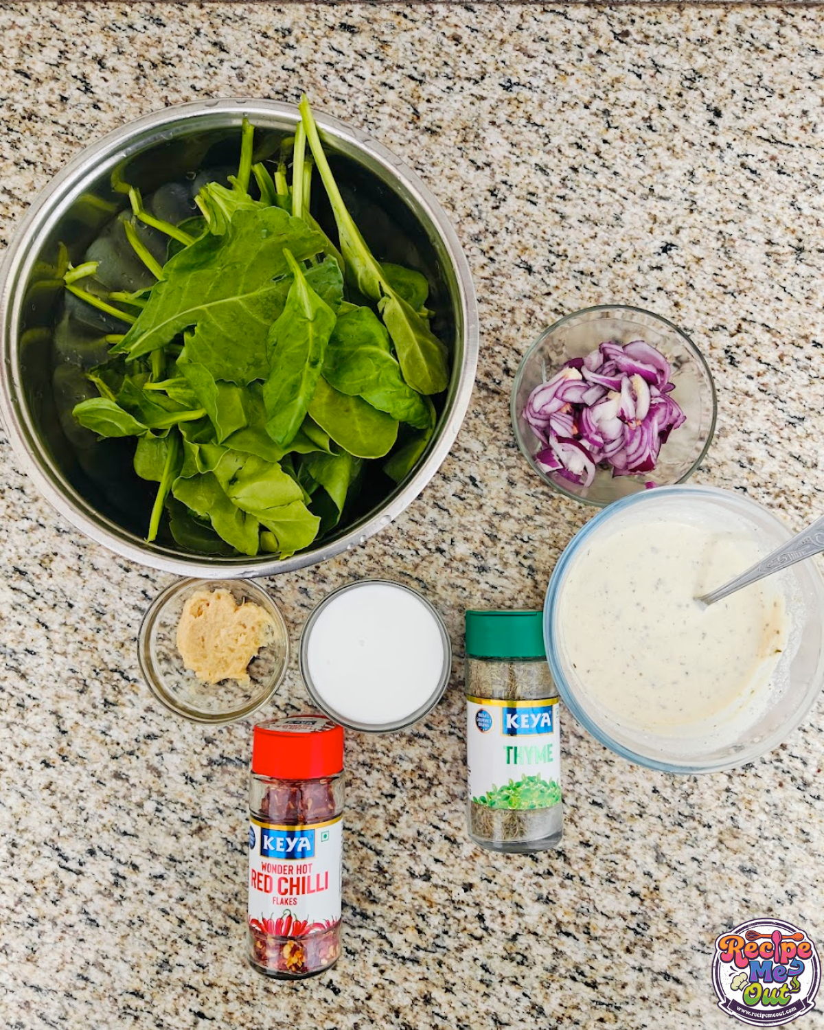 Ingredients for creamy spinach Alfredo displayed on a counter: fresh spinach leaves, chopped red onion, minced garlic, cream, coconut milk, alfredo sauce, chili flakes, Italian seasoning and more. 